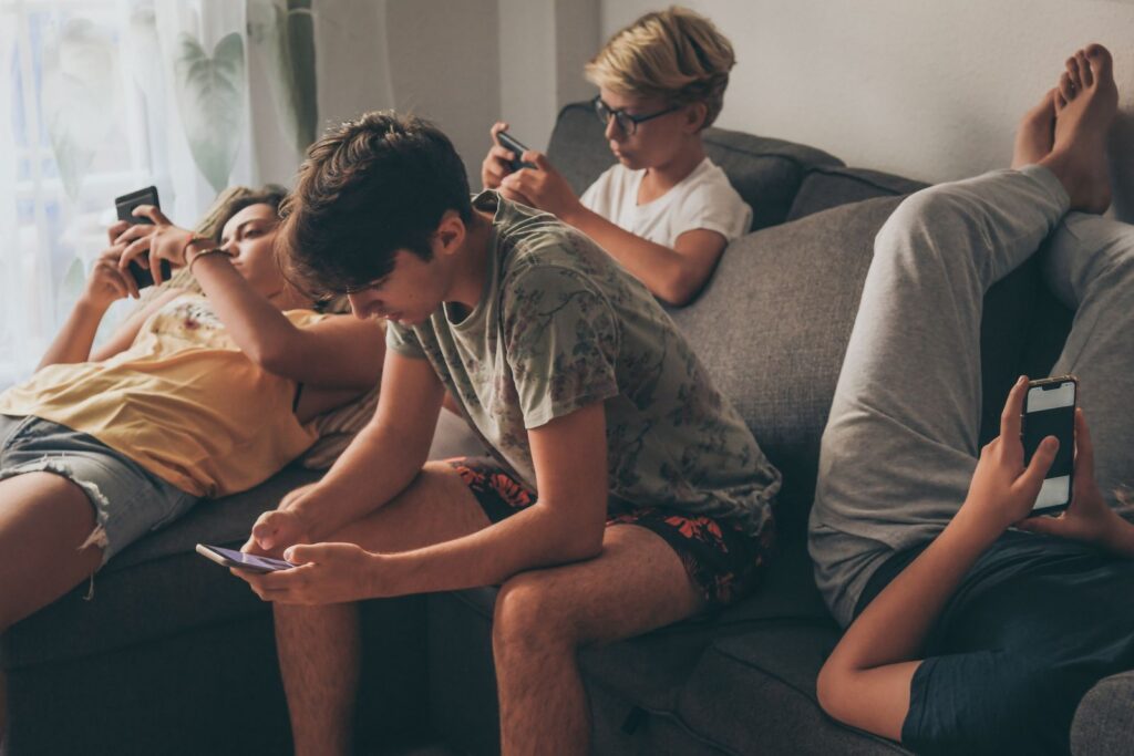 Group of teenager using smartphone sitting on a sofa at home. Young boys and a girl sharing photo and video watching social story online. Friends enjoying new trend technology.